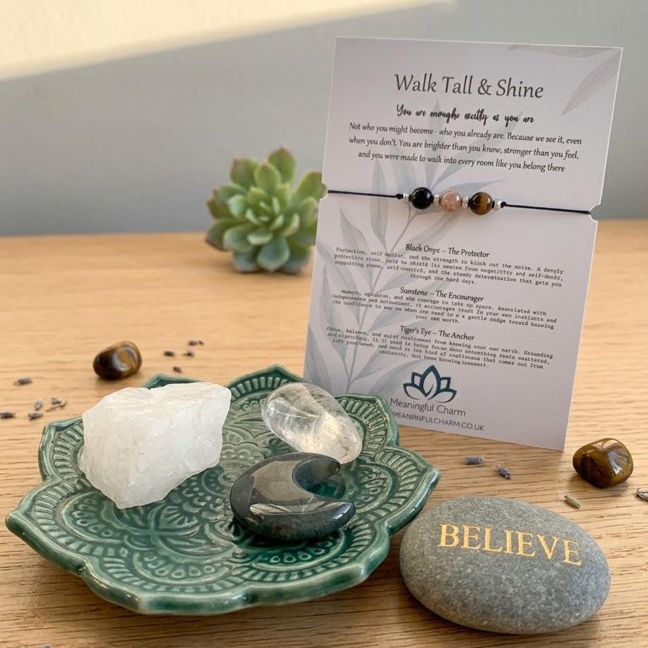 A beaded bracelet displayed on a printed card titled 'Walk Tall & Shine,' arranged on a wooden surface with crystals, a green ceramic dish, and a gray stone marked 'BELIEVE.'.
