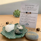 A beaded bracelet displayed on a printed card titled 'Walk Tall & Shine,' arranged on a wooden surface with crystals, a green ceramic dish, and a gray stone marked 'BELIEVE.'.