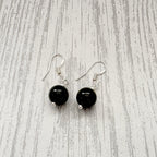 A pair of dangle earrings with round black beads and silver fishhook wires, displayed on a light gray wood-grain surface.