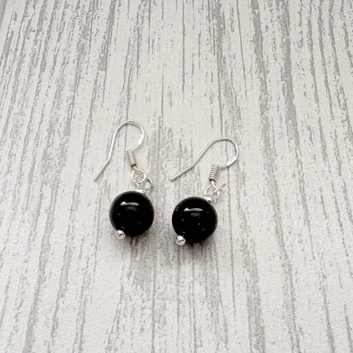 A pair of dangle earrings with round black beads and silver fishhook wires, displayed on a light gray wood-grain surface.