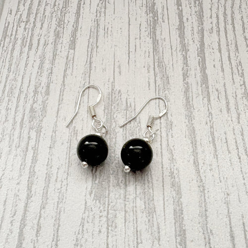 A pair of dangle earrings with round black beads and silver fishhook wires, displayed on a light gray wood-grain surface.