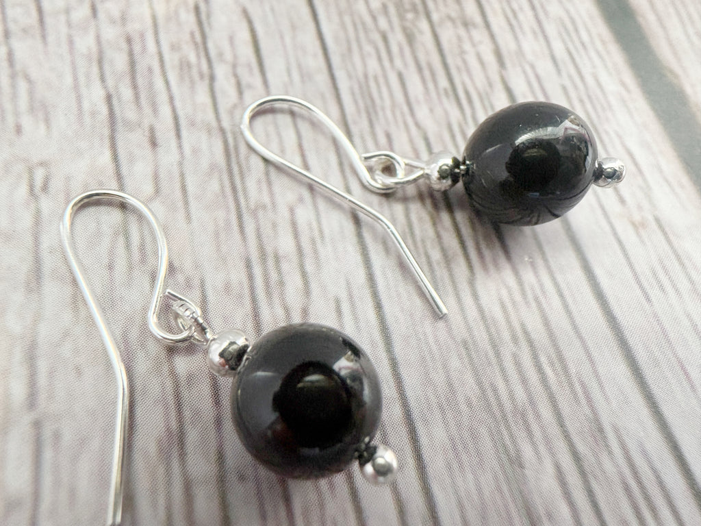 A pair of silver fishhook drop earrings with polished black spherical beads and small silver spacer beads, displayed on a light gray wood-textured background.