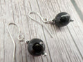 A pair of silver fishhook drop earrings with polished black spherical beads and small silver spacer beads, displayed on a light gray wood-textured background.