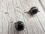 A pair of silver fishhook drop earrings with polished black spherical beads and small silver spacer beads, displayed on a light gray wood-textured background.