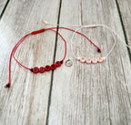 Two personalized cord bracelets displayed on a light wood background. One bracelet is red with red letter beads spelling 'BOBBY' and black accent beads, while the other is white with pink letter beads spelling 'PHIA' and gold letter accents.