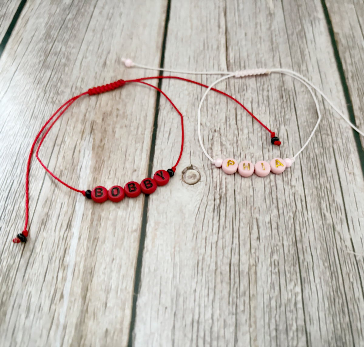 Two personalized cord bracelets displayed on a light wood background. One bracelet is red with red letter beads spelling 'BOBBY' and black accent beads, while the other is white with pink letter beads spelling 'PHIA' and gold letter accents.