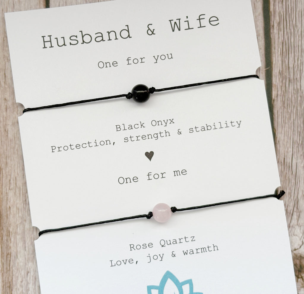 A gift set of two black cord bracelets on a white card, labeled 'Husband & Wife'. The top bracelet has a black onyx bead, and the bottom bracelet has a pale pink rose quartz bead.