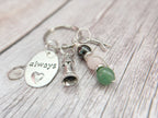 A close-up of a silver keychain with charms, including a round silver disc engraved with the word 'always' and a small heart, a silver tower charm, an X charm and a crystal bead stem with green, dark grey and pink beads, resting on a light wood background.