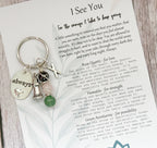 A personalised keychain with a silver-toned metal split ring and charms, placed on top of a printed card. The keychain includes a round silver charm engraved with the word 'always', a small silver tower, and a silver X Charms, and a crystal stem with green, pink and dark grey stone beads.
