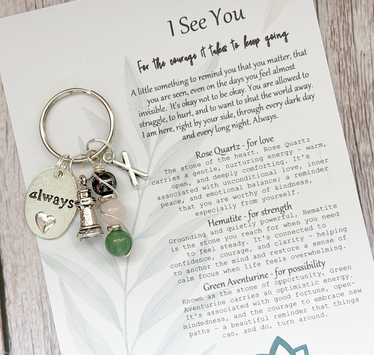 A personalised keychain with a silver-toned metal split ring and charms, placed on top of a printed card. The keychain includes a round silver charm engraved with the word 'always', a small silver tower, and a silver X Charms, and a crystal stem with green, pink and dark grey stone beads.