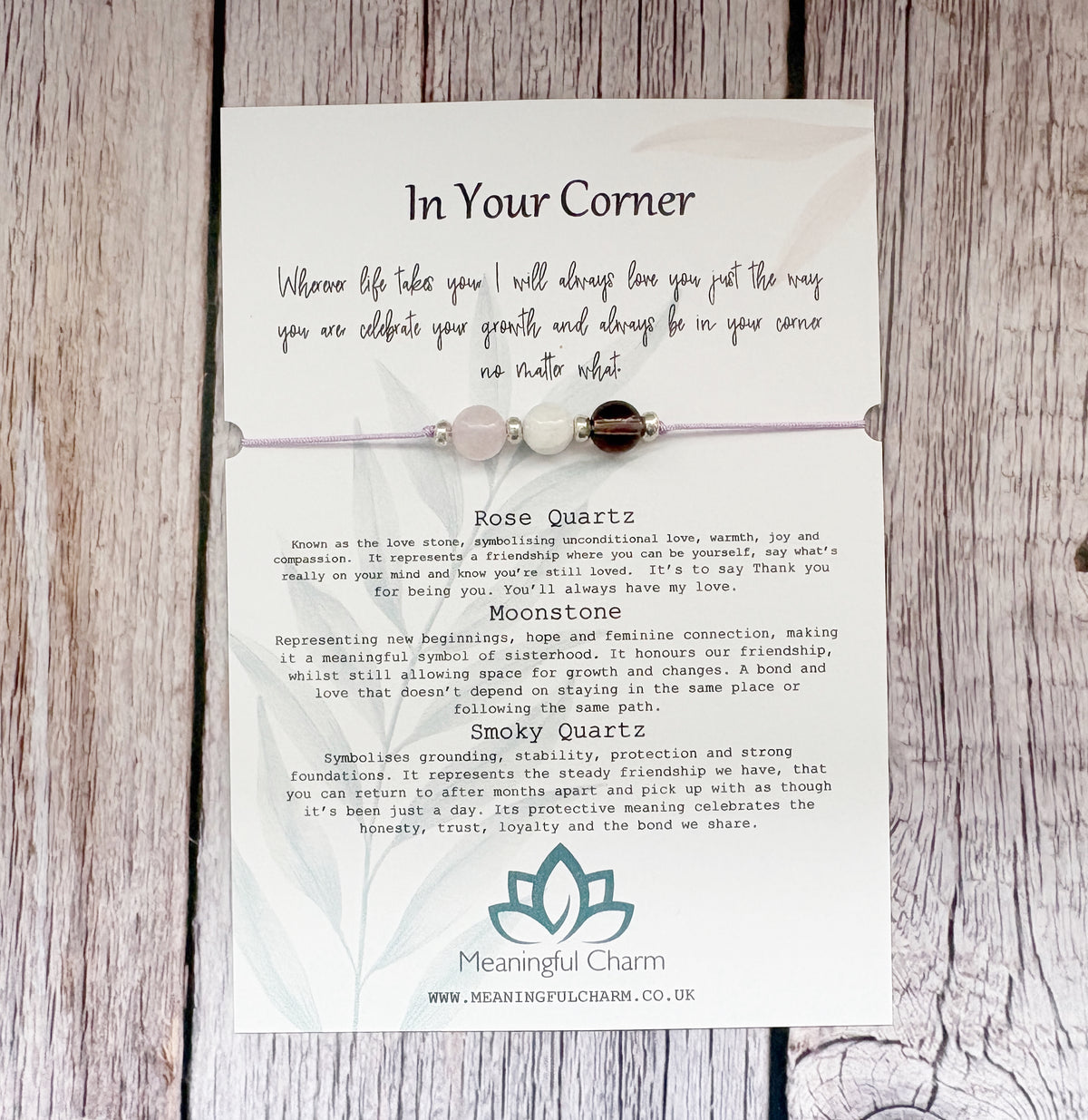 A square white card with printed text and a teal lotus logo, placed on a rustic whitewashed wooden surface. The card appears to be the product itself rather than an auxiliary chart, so this is Product/Packaging.