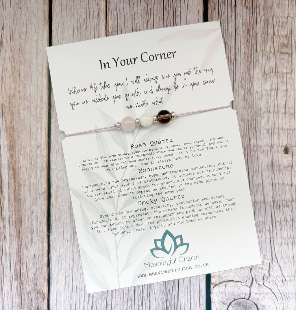 A delicate beaded bracelet card on a rustic wood background. The card features a rose quartz and moonstone bracelet with three beads, accompanied by printed text titled 'In Your Corner' and descriptions for Rose Quartz and Moonstone, branded by Mean Bright Charm.