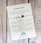 A delicate beaded bracelet card on a rustic wood background. The card features a rose quartz and moonstone bracelet with three beads, accompanied by printed text titled 'In Your Corner' and descriptions for Rose Quartz and Moonstone, branded by Mean Bright Charm.