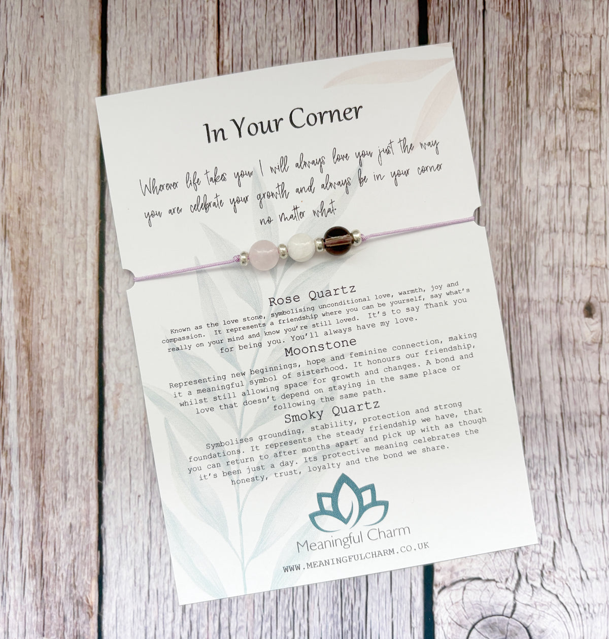 A delicate beaded bracelet card on a rustic wood background. The card features a rose quartz and moonstone bracelet with three beads, accompanied by printed text titled 'In Your Corner' and descriptions for Rose Quartz and Moonstone, branded by Mean Bright Charm.