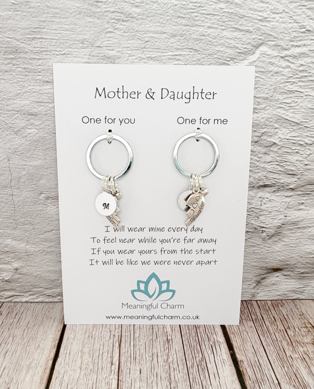 A pair of silver keychains with letter initial and crescent moon charms, presented on a white card labeled 'Mother & Daughter' by Meaningful Charm, set against a light gray textured wall and wooden floor background.