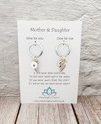A pair of silver keychains with letter initial and crescent moon charms, presented on a white card labeled 'Mother & Daughter' by Meaningful Charm, set against a light gray textured wall and wooden floor background.