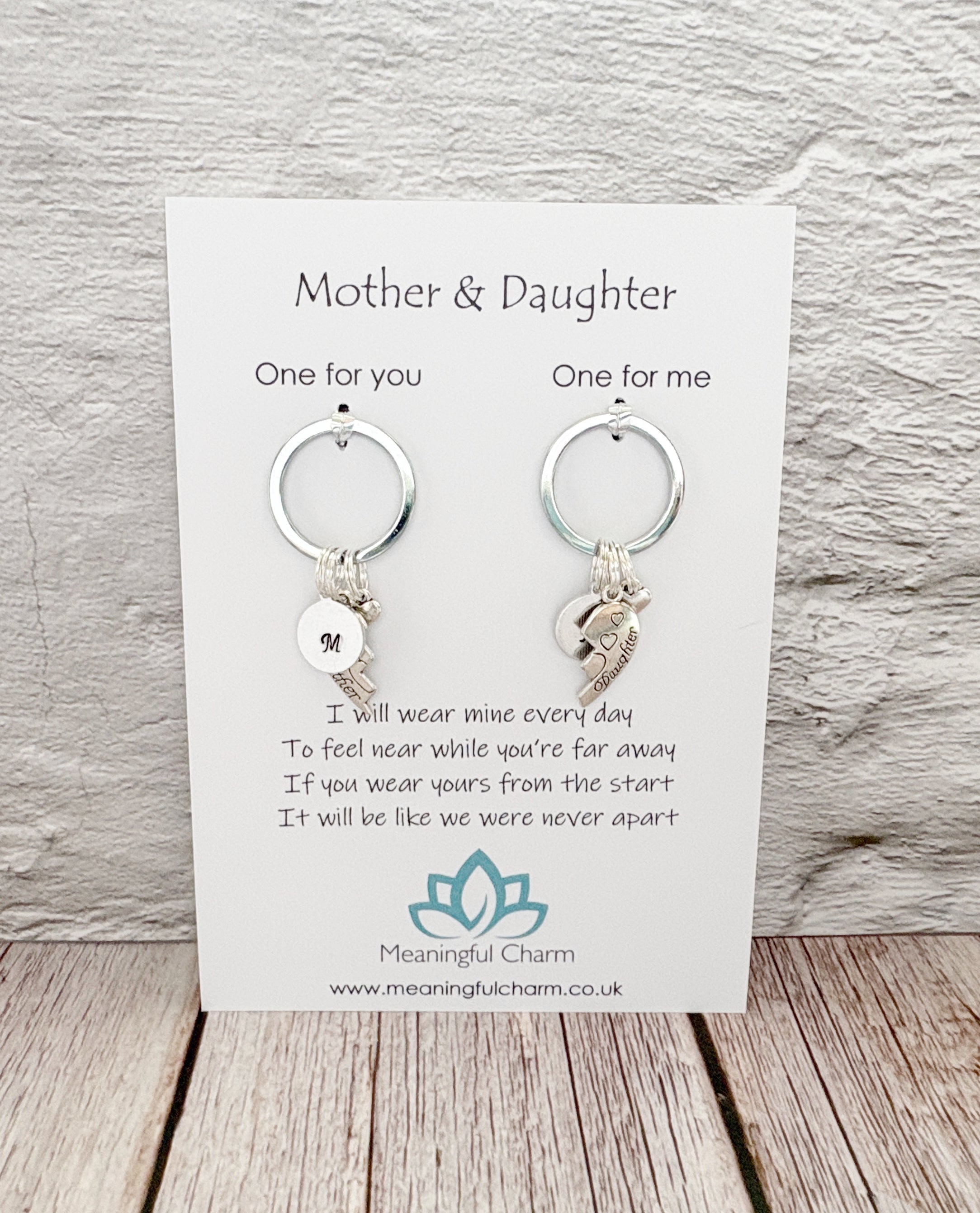 A pair of silver keychains with letter initial and crescent moon charms, presented on a white card labeled 'Mother & Daughter' by Meaningful Charm, set against a light gray textured wall and wooden floor background.