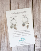 A pair of silver keychain earrings with bird charms and initial plates, displayed on a white card labeled 'Mother & Daughter' on a rustic wooden background. The card includes a quote and the brand 'Meaningful Charm'.