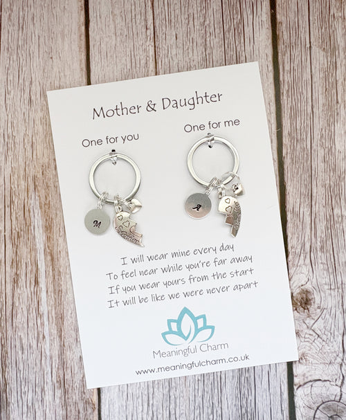 A pair of silver keychain earrings with bird charms and initial plates, displayed on a white card labeled 'Mother & Daughter' on a rustic wooden background. The card includes a quote and the brand 'Meaningful Charm'.