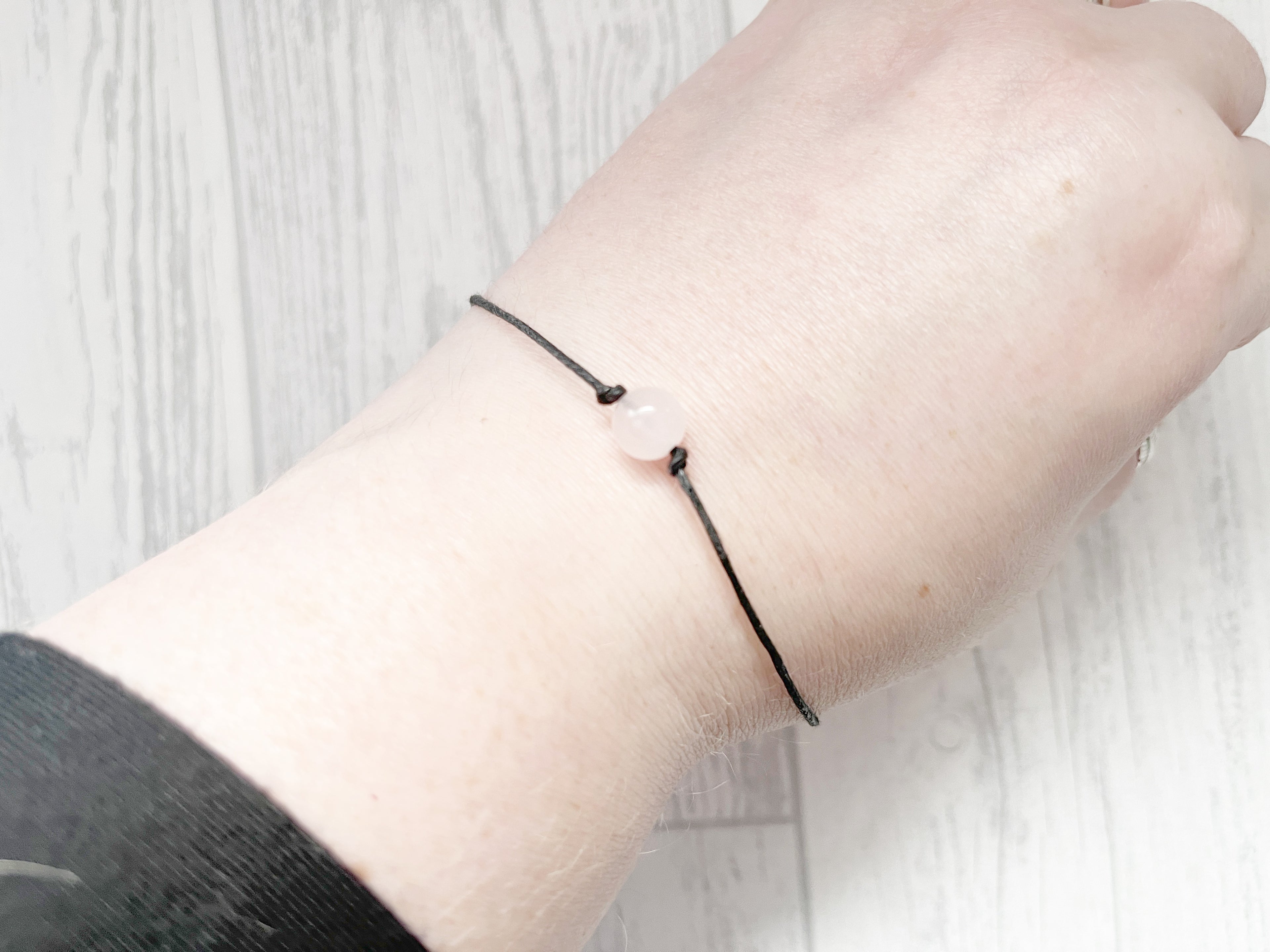 Close-up of a wrist wearing a black bracelet with a pink bead on a light wooden surface.