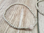 A beaded stretch bracelet featuring small silver metallic beads and one round pale pink stone bead, displayed on a light wood grain surface.