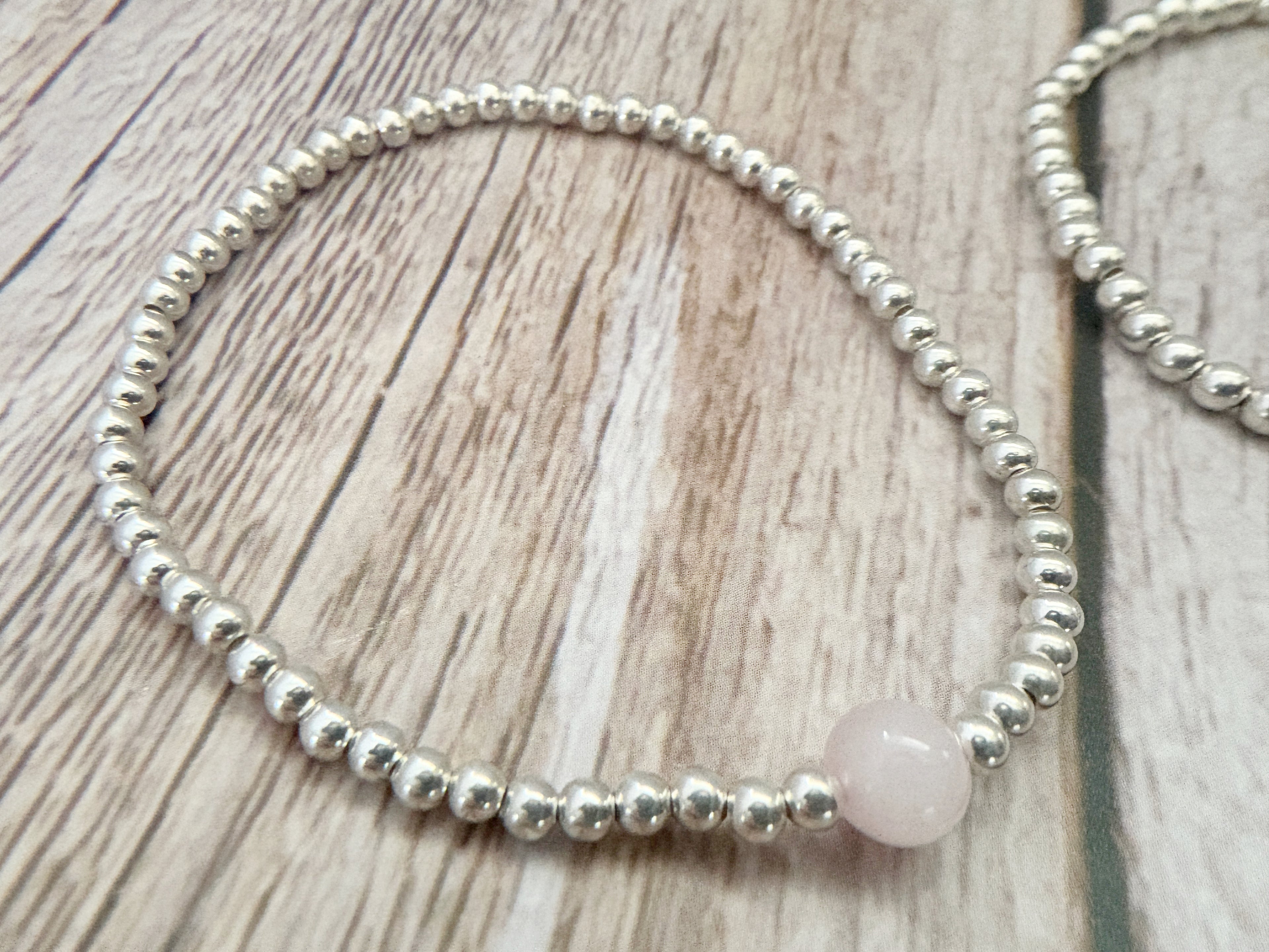 A beaded stretch bracelet featuring small silver metallic beads and one round pale pink stone bead, displayed on a light wood grain surface.