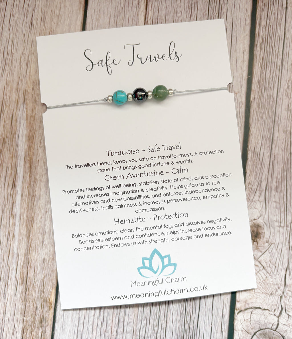 A beaded cord charm labeled 'Safe Travels' by Meaningful Charm, featuring gemstone beads, turquoise, hematite and green aventurine on a white card with text, displayed on a rustic wooden surface.