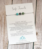 A beaded cord charm labeled 'Safe Travels' by Meaningful Charm, featuring gemstone beads, turquoise, hematite and green aventurine on a white card with text, displayed on a rustic wooden surface.