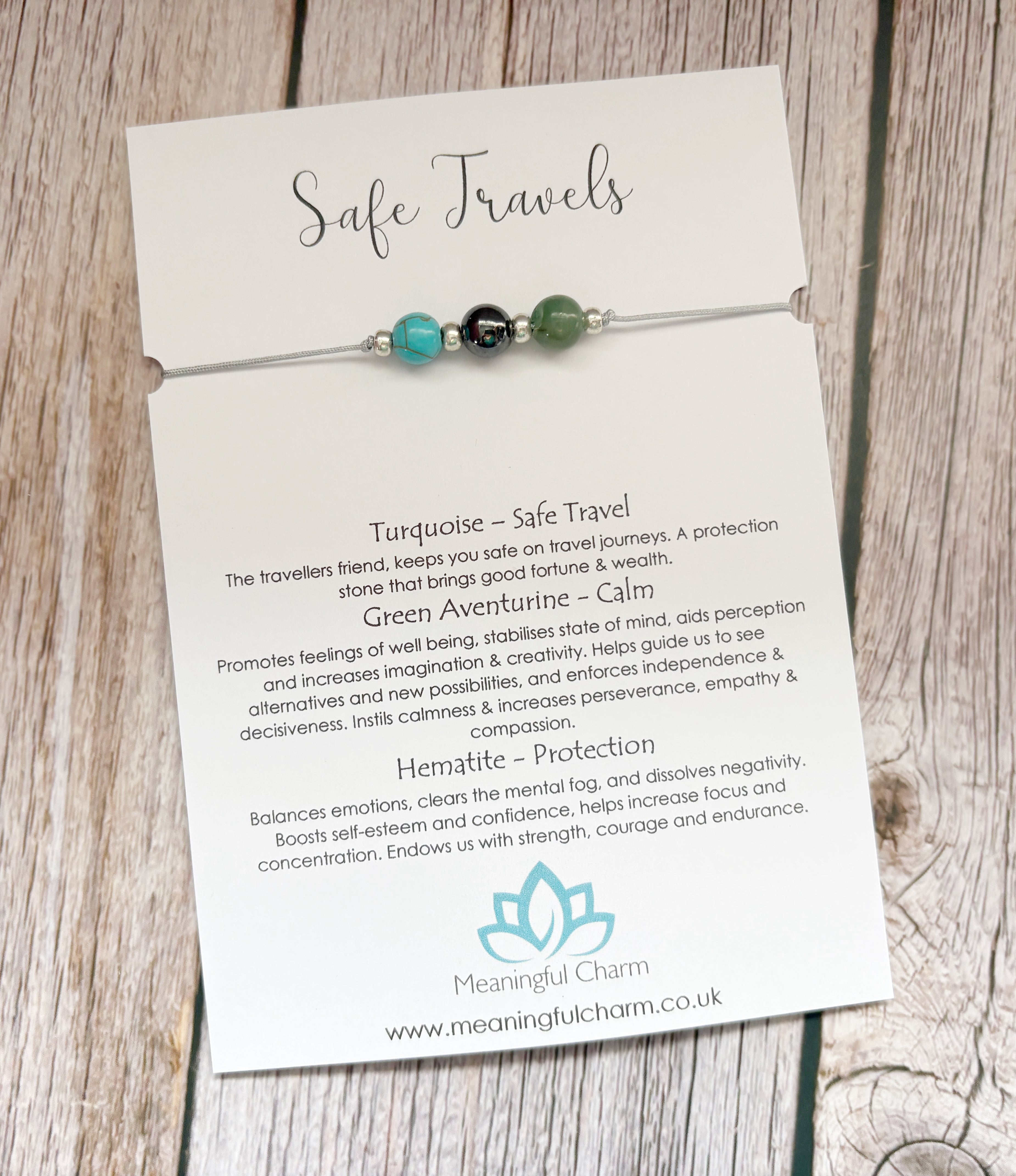A beaded cord charm labeled 'Safe Travels' by Meaningful Charm, featuring gemstone beads, turquoise, hematite and green aventurine on a white card with text, displayed on a rustic wooden surface.