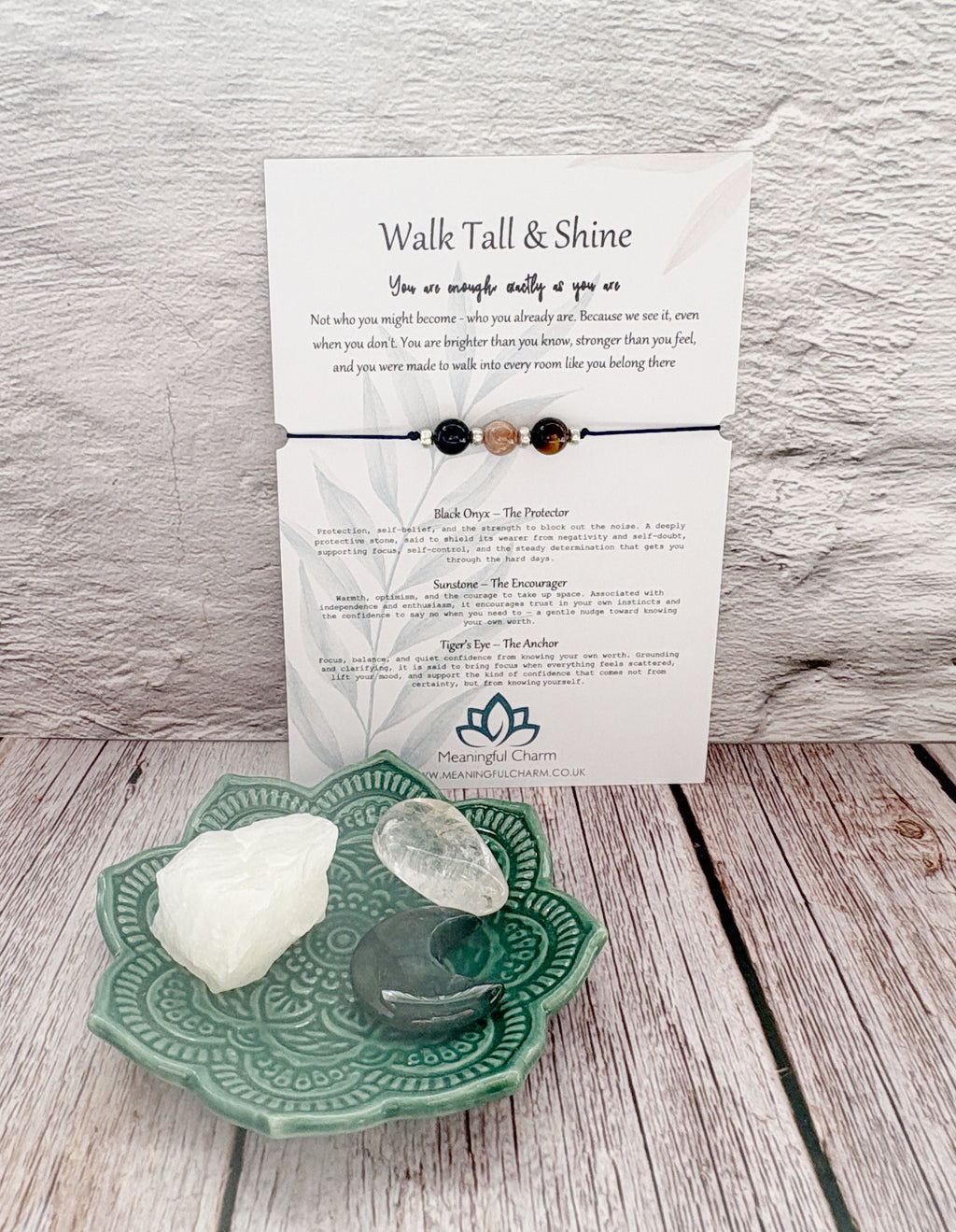 A beaded affirmation bracelet with four dark beads is displayed on a branded card that reads 'Walk Tall & Shine,' alongside two crystals on a decorative teal plate against a rustic wood and white wall background.