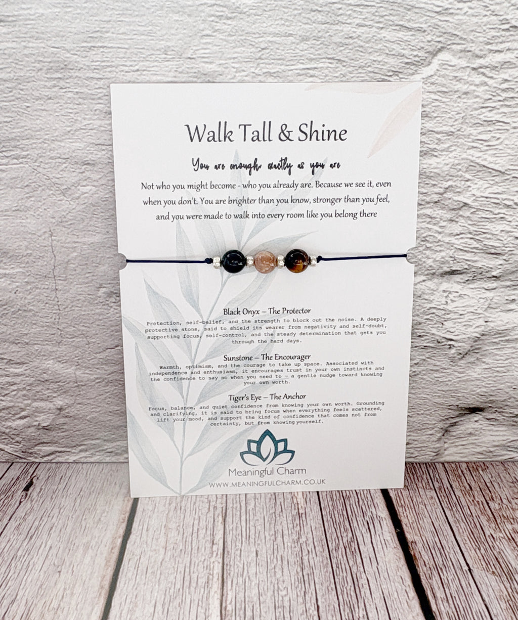 A white card with printed affirmation text and three small gemstone beads on a thin cord, displayed against a light wood plank background. The card includes printed meanings for Black Opal, The Protector, and Sweetheart.