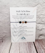 A white card with printed affirmation text and three small gemstone beads on a thin cord, displayed against a light wood plank background. The card includes printed meanings for Black Opal, The Protector, and Sweetheart.
