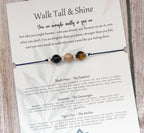 A printed card or booklet page titled 'Walk Tall & Shine' with inspirational text, draped over a silver chain with a small amber bead, on a light wood background.