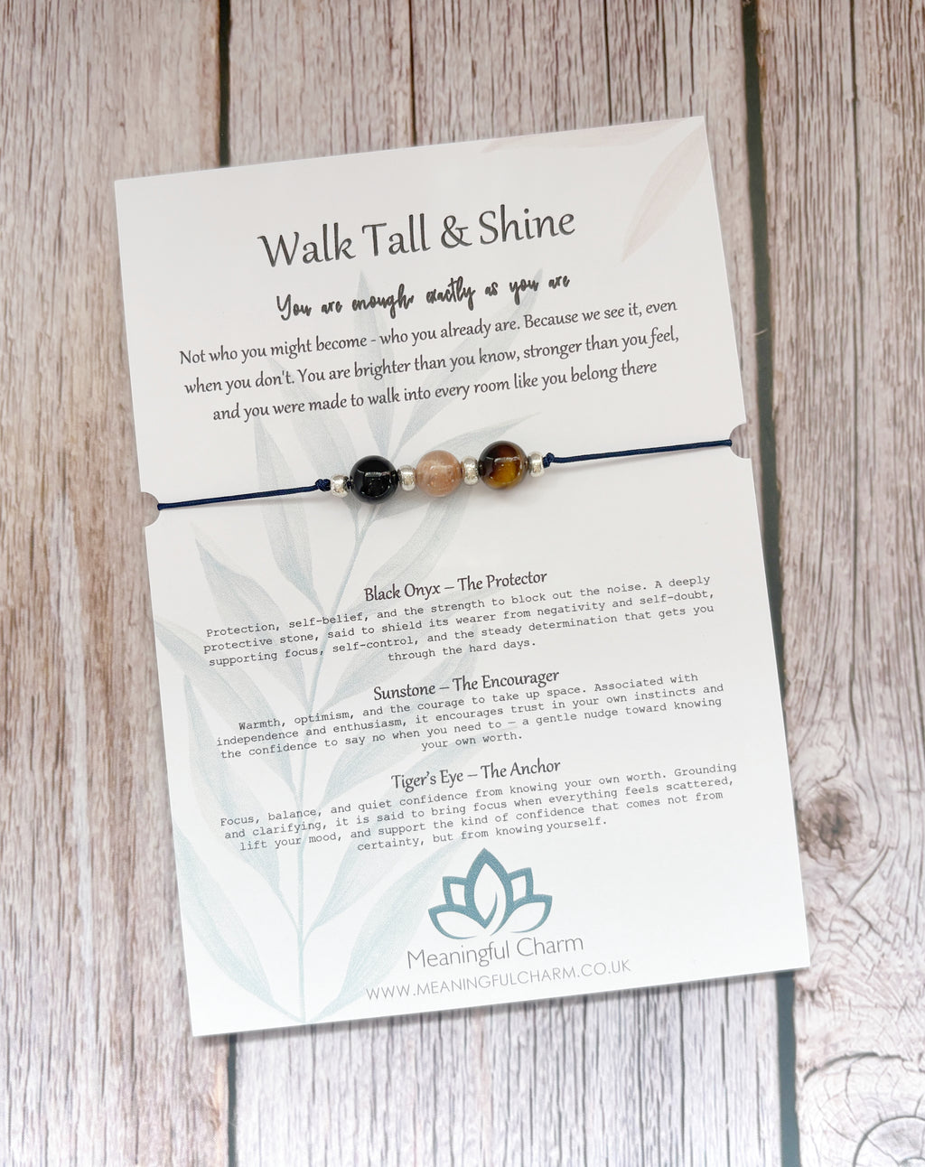 A beaded black cord bracelet with three beads (black, brown, and cream) displayed on its printed backing card, which features inspirational quotes and product descriptions for 'Meaningful Charm'.