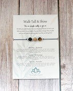 A beaded spiritual necklace with black, white, and brown beads displayed on its printed backing card, which reads 'Walk Tall & Shine' and features descriptions for the beads and meanings, set against a light wood background.