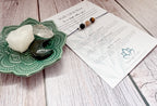 A beaded spiritual cord bracelet with black, white, brown, and tiger eye beads, displayed on its printed packaging card next to a green ceramic dish holding crystals, all on a whitewashed wooden surface.